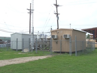 Two small metal buildings with a platform between them containing air monitoring equipment in a grassy parking lot, with a power line behind the buildings.