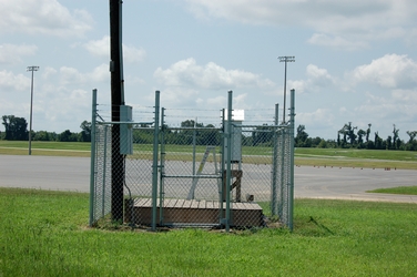 A fenced in area holding a wooden platform with a piece of air monitoring equipment, with a parking lot and trees in the background.