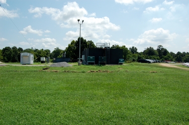 A grassy field with a large, black, metal industrial unit and a parking lot of cars to the right.