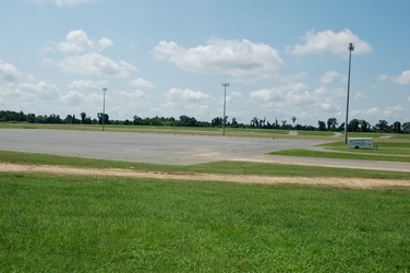 An empty, large concrete parking lot with grass surrounding and three light poles it and a small white trailer to the right.