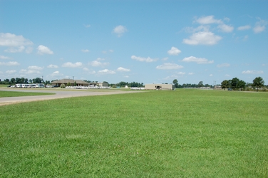 A large grassy field in the foreground with a concrete drive and parking lot with cars and a building to in the middle and to the left in the background.