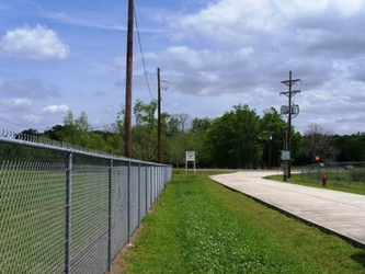 A chain length fence lining the left side of the picture, with grass in the middle and a concrete drive running lengthwise on the right.