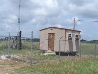 A tan metal building surrounded by chain-link fencing in the middle of a grassy field with cloudy skies overhead.