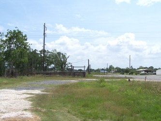 A field of grass with a gravel driveway, a parking lot in the distance, and wooden electrical poles and lines overhead.