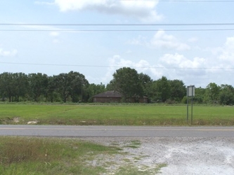 A road with a perpendicular gravel driveway entrance in the foreground, and a grass field with trees in the background.