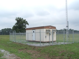 A tan metal building on a gravel pad inside a fenced-in area in the middle of a grass field.