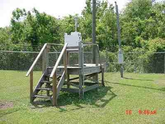 A wooden platform, accessible by stairs, that holds an FRM air sampling instrument in the middle of a grass field with a chain-link fence and trees running along the background.