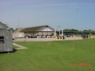 A grassy yard with a shed to the left and a large building in the background with cars parked under an overhang within a parking lot.