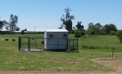 A grass and dirt field with a small shed inside a chain-link fenced-in square with more grass and trees in the background under a clear blue sky.