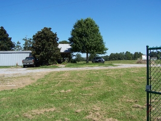 A field of grass and dirt with a gravel road in the near background and a building, cars, and trees in the further background under a clear blue sky.