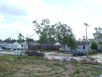A neighborhood street with a grass field in the foreground and houses in the background. There are trees and a cloudy sky behind the houses.