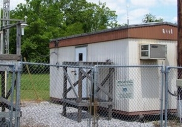 A tan metal building on a gravel pad with trees and blue skies in the background.