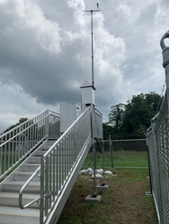 A metal staircase with a platform holding a piece of air monitoring equipment, large trees and a cloudy sky in the background.