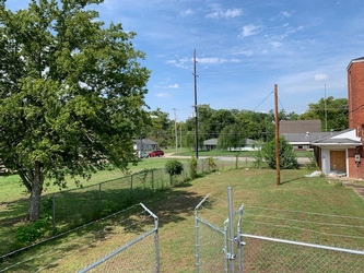 The corner of a chain link fence with an open gate with the side of a house to the right and a tree to the left.