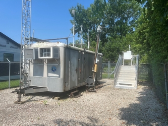 A fenced-in gravel plot with a camper trailer and meteorological tower on the left and a metal platform holding an air sampler on the right.