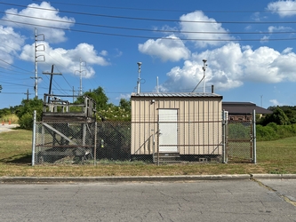 A small tan shed surrounded by a chain-link fence in the middle of a grass field. There is pavement in the foreground and a cloudy blue sky in the background.