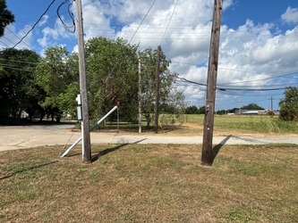 A road in between fields with electrical poles lining the road. There is a cloudy blue sky and electrical lines overhead.