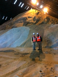 Two inspectors in orange safety vests standing in front of large piles of sugar while examining equipment inside a sugar mill