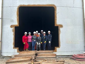 Eight inspectors wearing safety gear standing together in the large opening of a massive industrial tank inside a facility