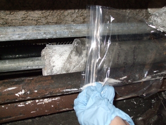 Blue‑gloved hand holding plastic wrap close to asbestos insulation on a pipe