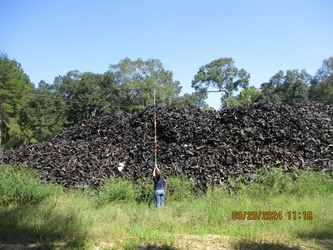 Large pile of waste tires in front of an overgrown wooded area, with an inspector holding a stick about ten feet in the air to take a measurement.