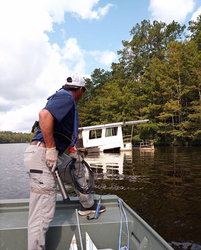 Male inspector standing with one foot on bow of deck using steel container to take sample from river. 