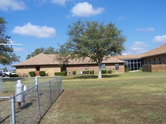 A grassy yard with a chain link fence, brick buildings, and trees.