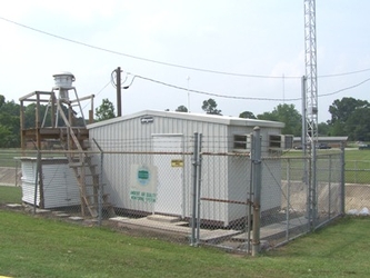 A small white building and attached wooden platform on a dirt plot inside a chain-link fenced square surrounded by short grass.