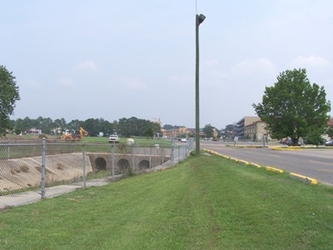 A clear grass strip extending vertically down the middle with a concrete ditch and culverts on the left, delineated by chain-link fencing, and a road on the right.