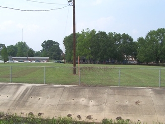 A concrete ditch in the foreground and a vast grass field behind it, separated by a chain-link fence barrier, with an electrical pole in the center of the field and trees in the background.