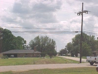 A road passing horizontally in the foreground with a house, trees, and cloudy sky in the background.