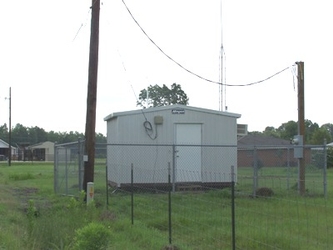 A small building inside a fenced-in grassy area with electrical poles nearby and trees in the far background.