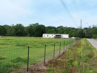 A large grassy field to the left, bordered by a fence with a ditch and road on the right side; there is a mobile home and trees in the background.
