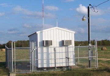 A small white building inside a chain-link-fenced, grassy area. There is an electrical pole and a cloudy sky in the background.