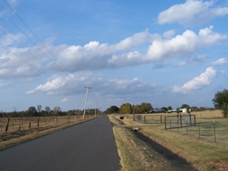 A road on the left side extending toward the horizon, flanked by fenced-in grass fields with a cloudy sky overhead.