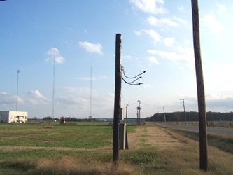 A field of short grass and dry dirt with a road to the right, a small white building on the far left, and electrical poles scattered throughout the view.