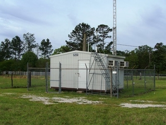 A small building in the center of a fenced-in section of a wide grassy field with trees in the background.