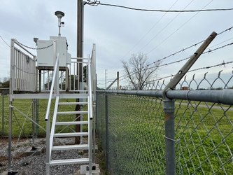 A metal staircase on the left, leading up to a metal platform holding a piece of air monitoring equipment. There is a chain-link fence to the right and a field in the background.