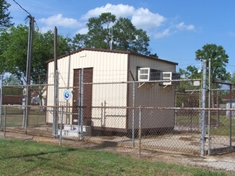 A small shed inside a chain-link fenced area with no grass. There is grass in the foreground outside of the fence and trees in the background under a blue sky.