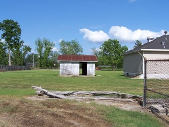 A grass and dirt field with wooden debris in the foreground, a shed in the center, trees in the far background, and part of chain-link fencing and a house partly visible to the right.