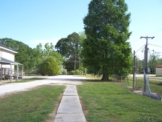 A paved sidewalk leading to a gravel lot with grass on either side. There is a building partly visible to the left and trees in the background.