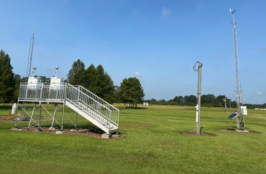 A grass field with a metal staircase and platform on the left holding two FRM air sampling instruments. There are some metal towers in the foreground and trees in the background.