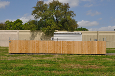 A solid wooden fence running horizontally through the middle of a grass yard with the top of a shed visible on the other side of the fence, as well as a concrete barrier and trees in the background.