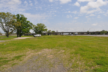 An empty grass and dirt field near an interstate highway to the right with a truck, trees, and a bridge in the background.