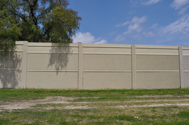 A tall, solid concrete barrier with some grass in the foreground and the top of a tree hanging over the fence.