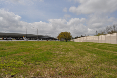 An expansive field of short grass and dirt with a highway and bridge to the left, a solid concrete wall on the right, and trees in the background.