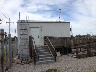 A small shed with stairs, a ladder, and an accessible ramp leading to the front. The shed sits in a fenced-in gravel plot under a cloudy sky.