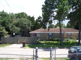 A house in a residential neighborhood with a road and chain-link fence in the foreground and trees in the background.