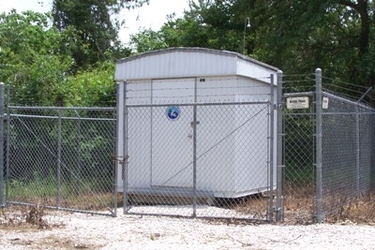 A small white shed inside a square of chain-link fencing on a plot of gravel and dead grass in front of a wooded background.