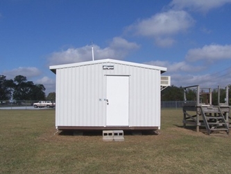 A white shed in a field with a wooden platform to the right and a cloudy blue sky overhead.